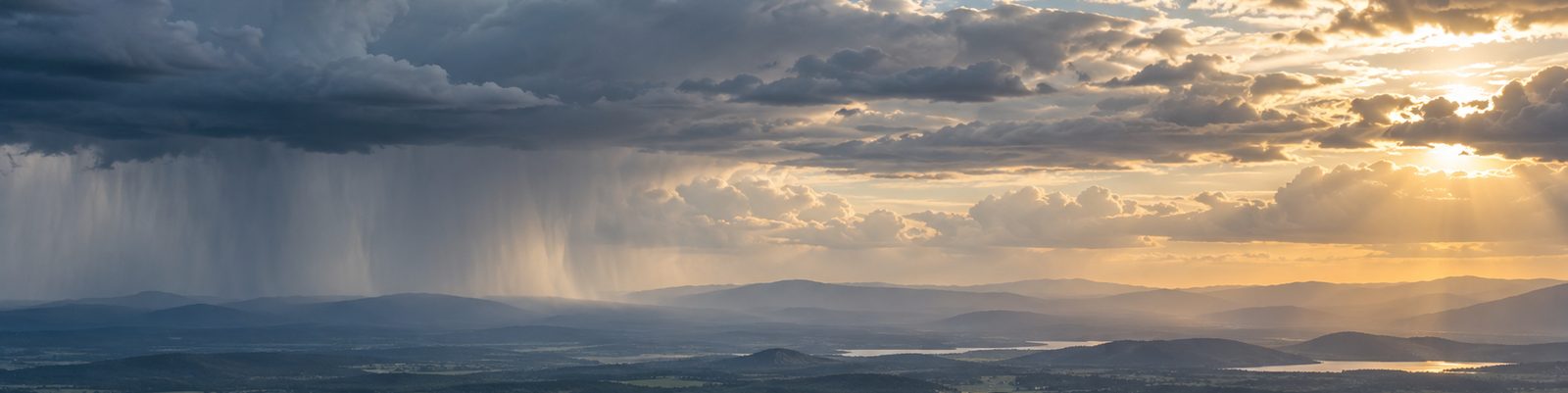 A wide evening scene in which rain falls from a dark cloud base on one side of the frame while broken cloud and low sunlight open over distant ridges on the other.